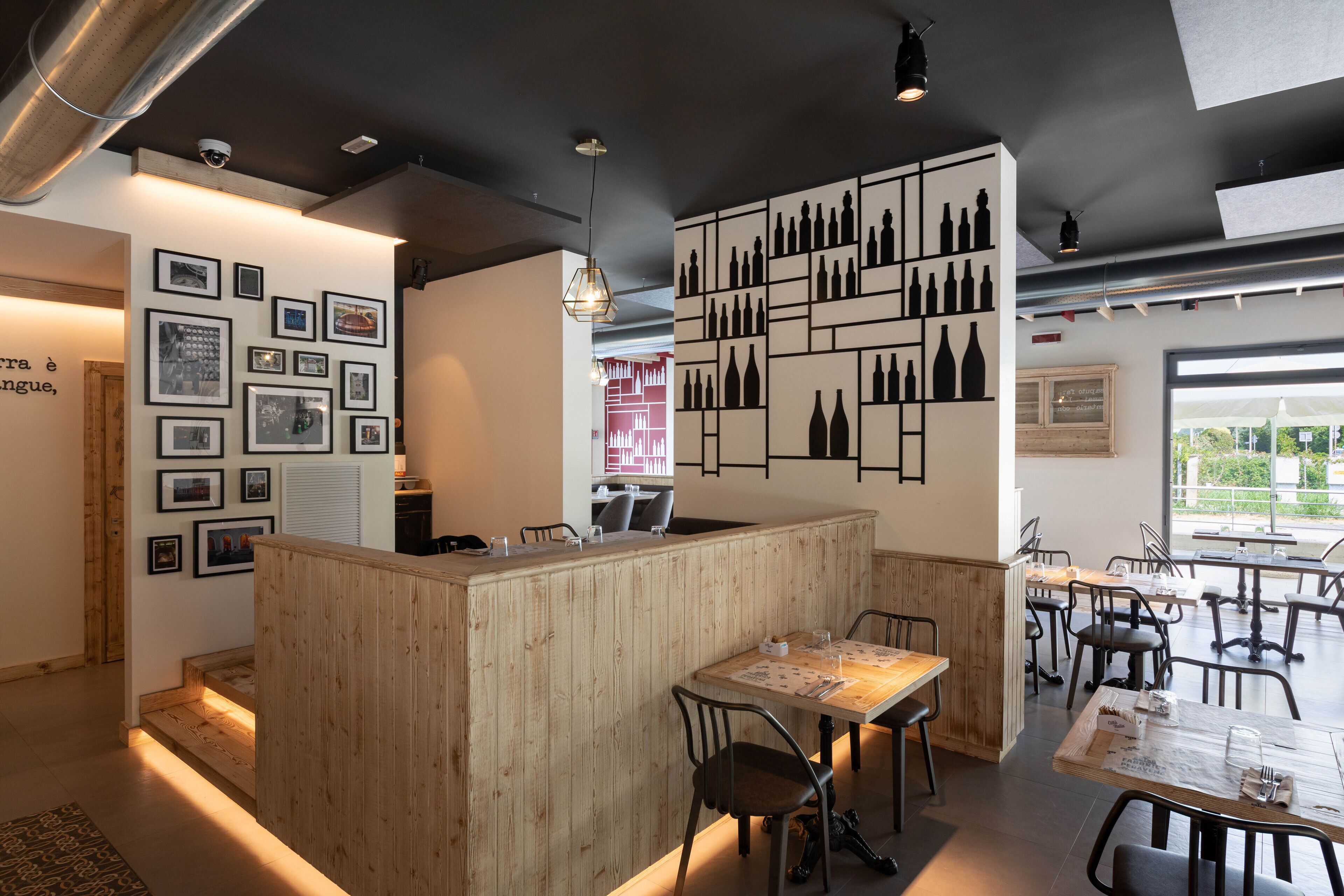 Un restaurant avec un comptoir en bois, plusieurs tables et des chaises. Les murs sont ornés de tableaux encadrés et une grande étagère expose de nombreuses bouteilles de vin.