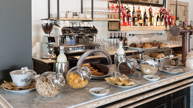 Una barra de desayuno con diversos alimentos, una máquina de café y una selección de botellas de vino.