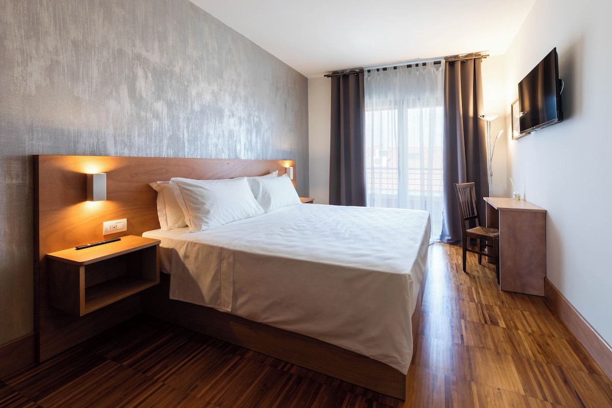 A hotel room with a wooden bed, bedside table, lamp, and a flat-screen TV mounted on the wall.