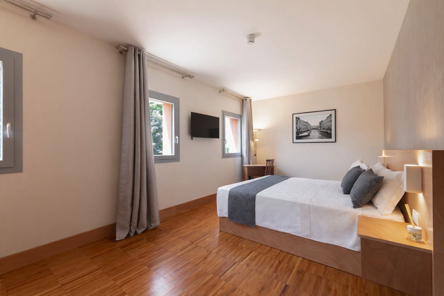 A bedroom with a bed, a desk, a chair, a TV, and a window.