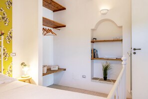 A bedroom with a bed, a nightstand, a lamp, a wall-mounted shelf with books and a plant, and a door leading to another room.