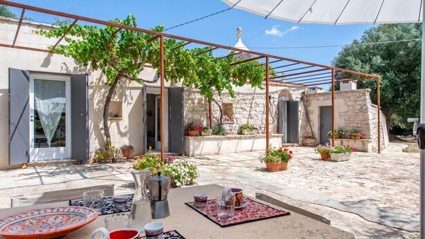 Une terrasse extérieure avec une table dressée pour le repas, entourée de plantes en pot et agrémentée d’une pergola.
