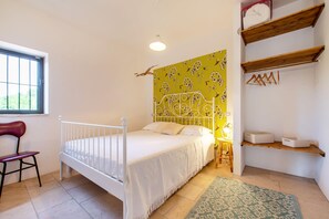 Une chambre avec un lit blanc, une chaise rouge, une fenêtre, un mur jaune recouvert d’un papier peint à motifs floraux et des étagères en bois.