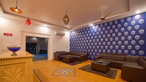 A living room with a brown sofa set, a coffee table, and a patterned rug. There is a blue wall with a floral design and a ceiling fan. - Desert Drop (Jaisalmer)
