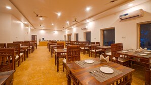 A spacious dining hall with multiple wooden tables and chairs, ceiling fans, and an air conditioning unit. - Desert Drop (Jaisalmer)