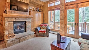 A stone fireplace with a television above it, a leather armchair with a red and beige throw blanket, and a wooden deck visible through large glass doors.