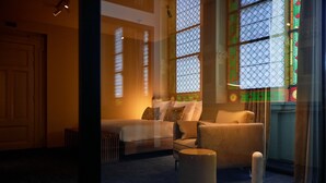 A cozy room with a sofa, ottoman, and a large window with stained glass. - Heavens Hotel (Hoorn)