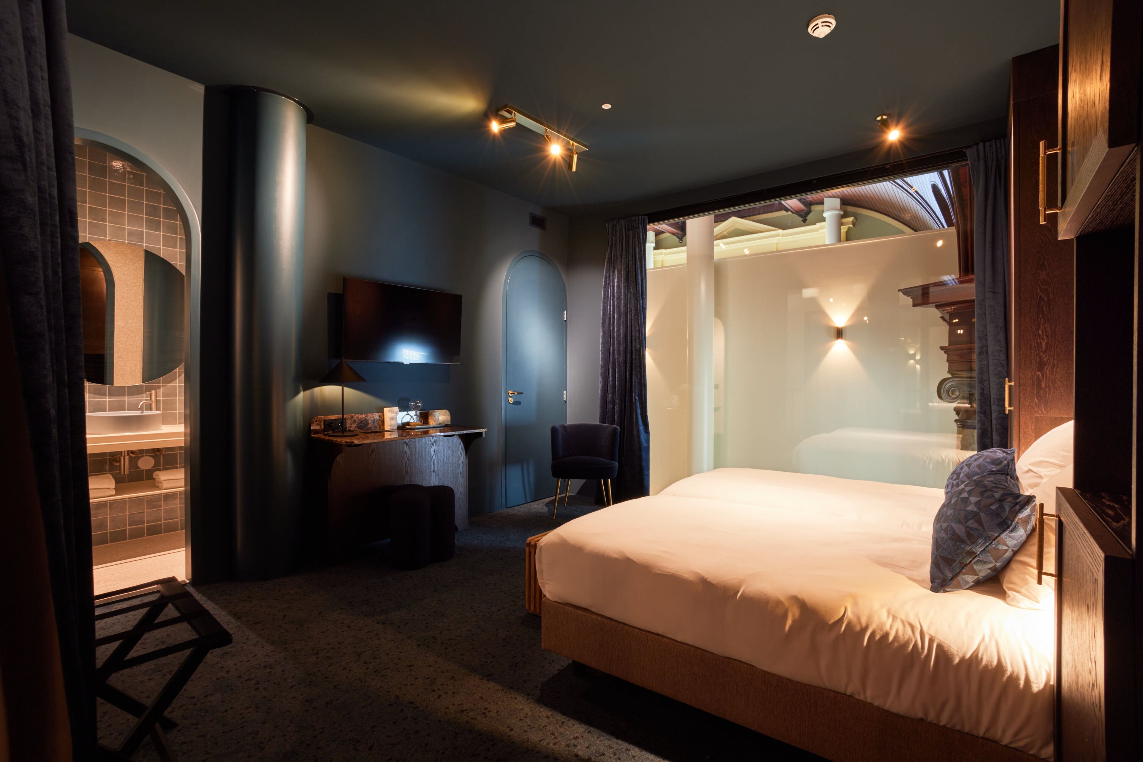 Una habitación de hotel moderna con una cama grande, un escritorio con una silla, un televisor y un baño visible a través de una puerta de cristal.