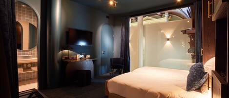 Una habitación de hotel moderna con una cama grande, un escritorio con una silla, un televisor y un baño visible a través de una puerta de cristal.