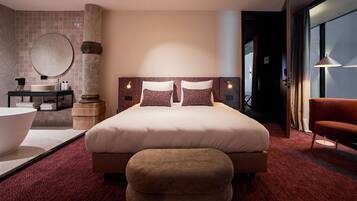 A modern hotel room with a large bed, a round mirror, a bathtub, and a seating area.