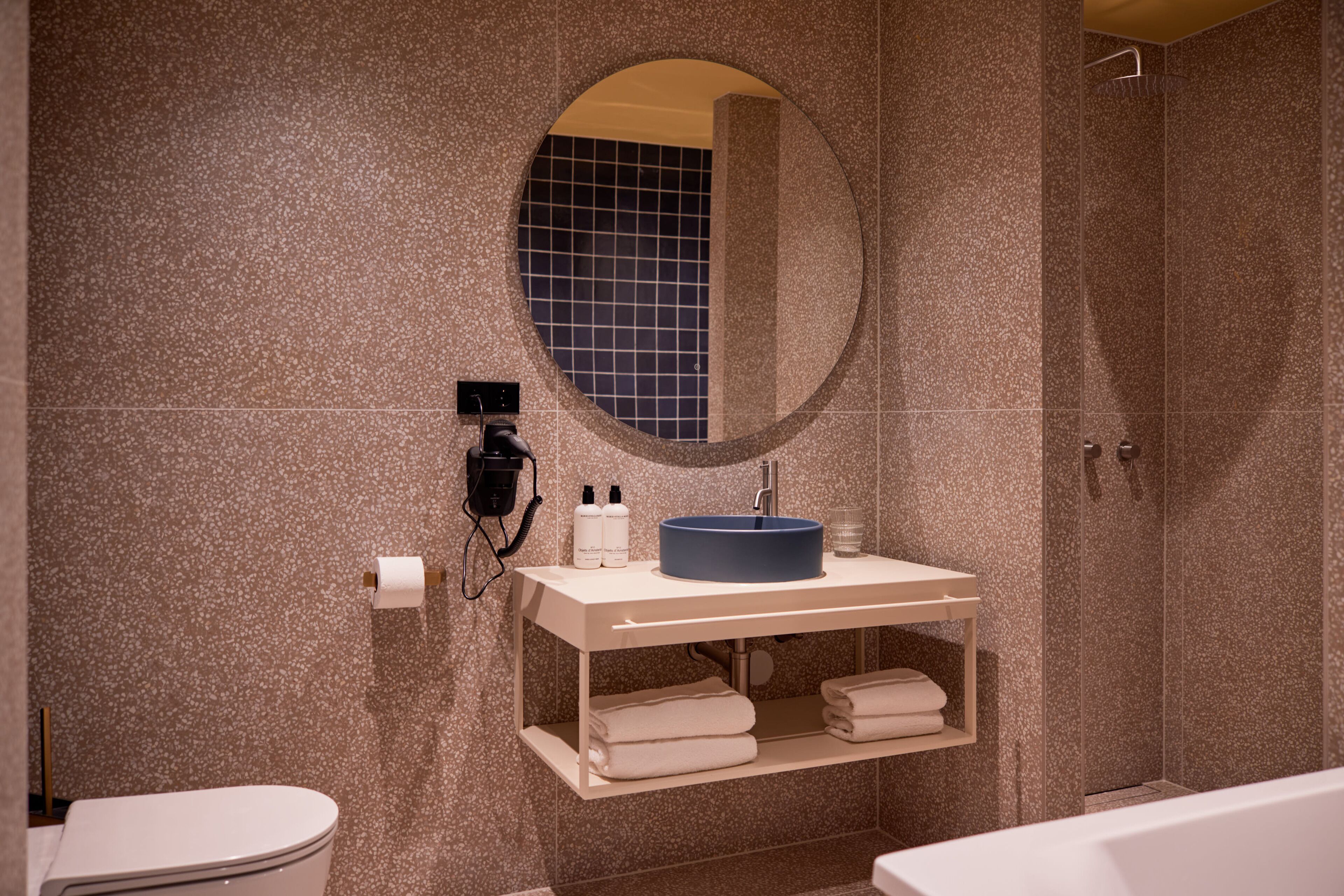 Une salle de bain avec un miroir rond, un lavabo avec un receveur bleu, des toilettes et un espace douche.