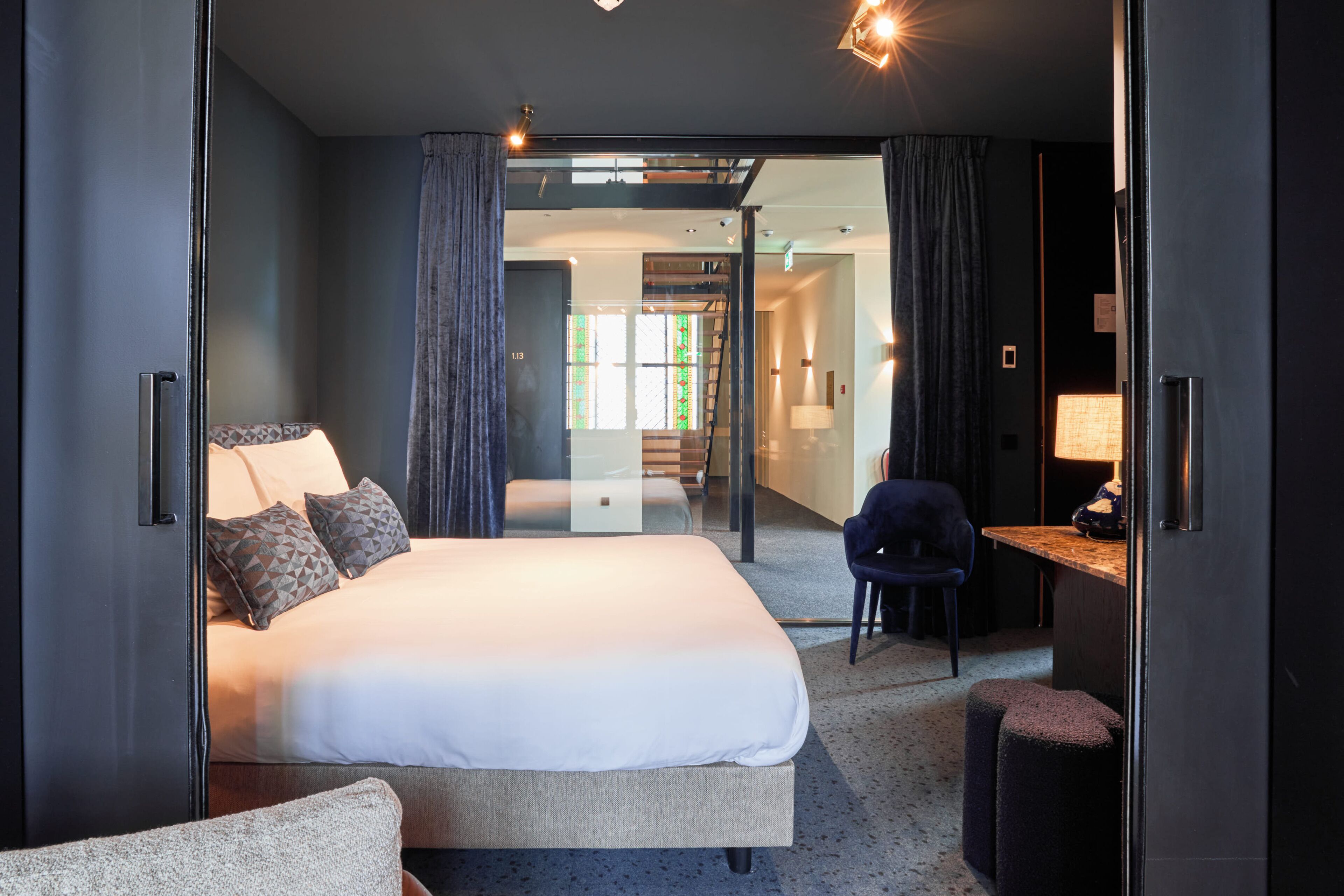 A modern hotel room with a large bed, a desk, and a chair. There is a glass partition separating the room from the hallway.