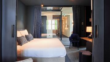 A modern hotel room with a large bed, a desk, and a chair. There is a glass partition separating the room from the hallway.
