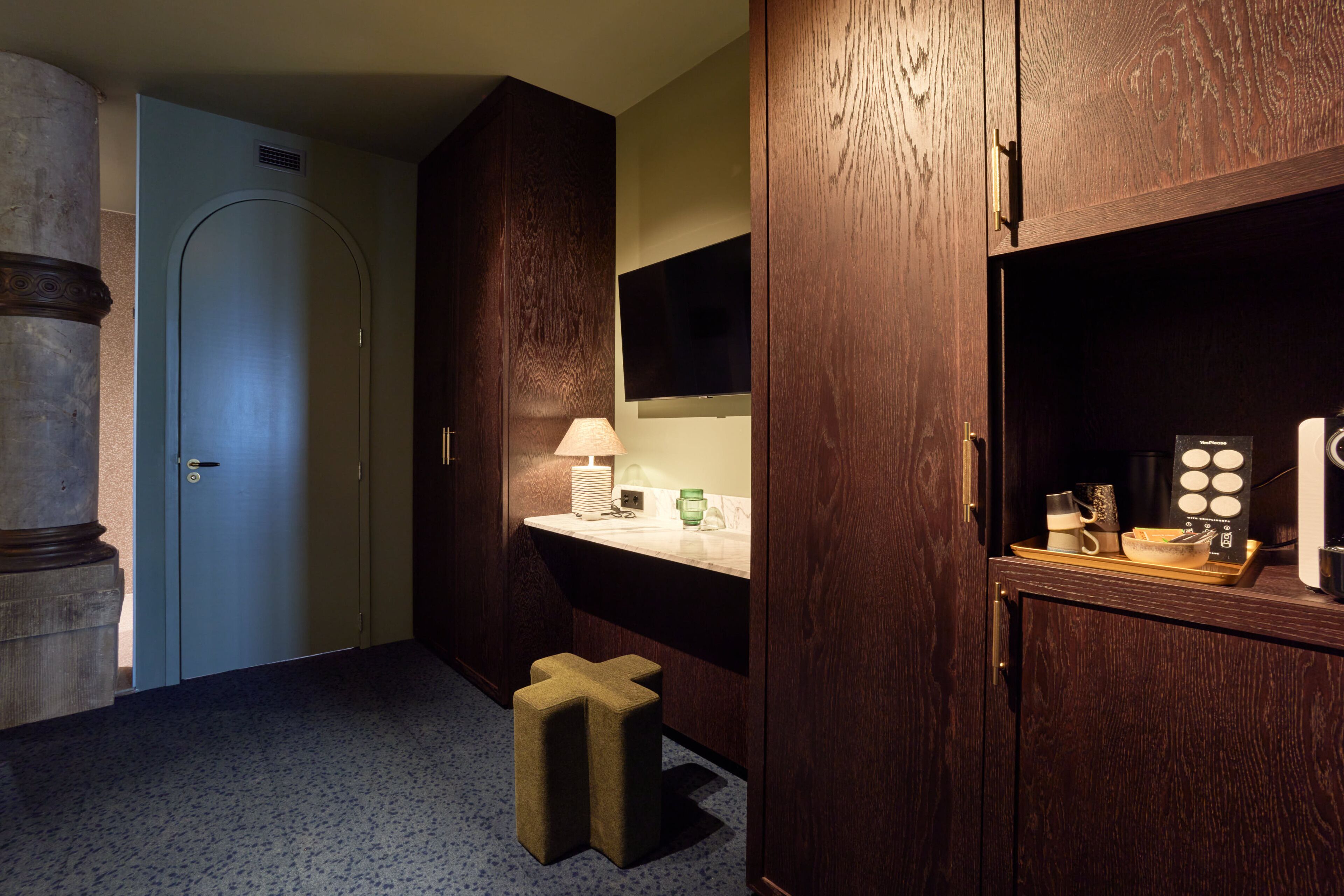 A hotel room with a wooden wardrobe, a vanity with a mirror, a small stool, and a tray with toiletries.