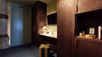 A hotel room with a wooden wardrobe, a vanity with a mirror, a small stool, and a tray with toiletries.