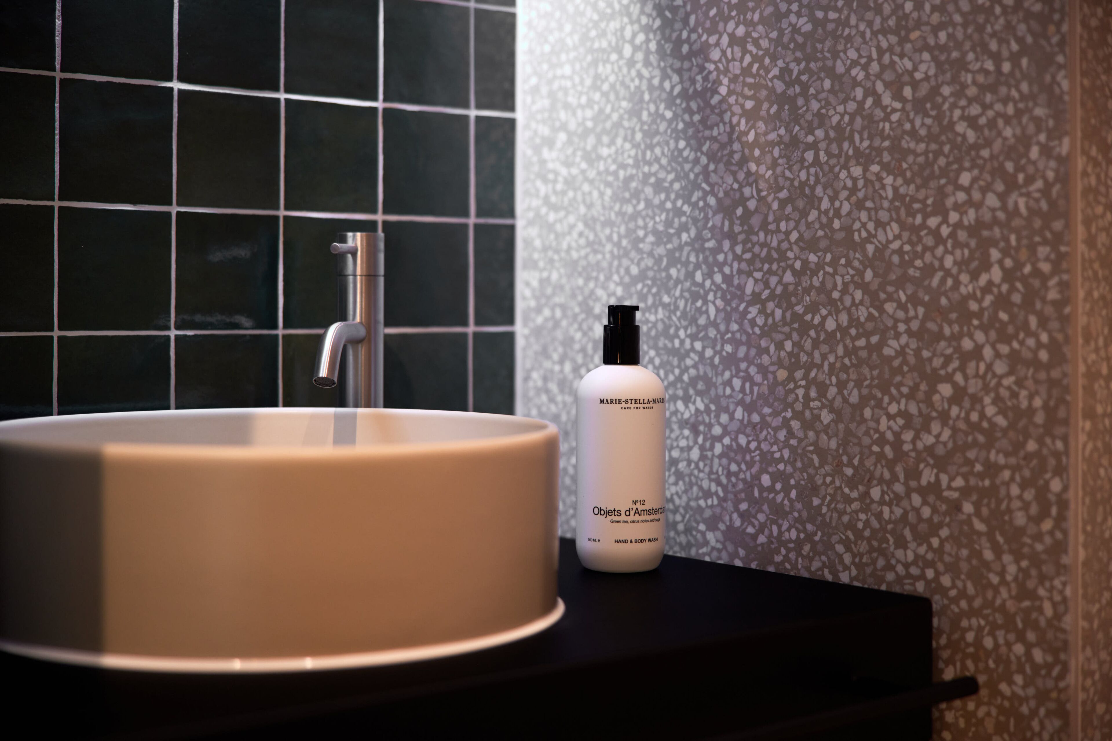 A bathroom with a white sink, a faucet, and a bottle of skincare product on a black countertop.