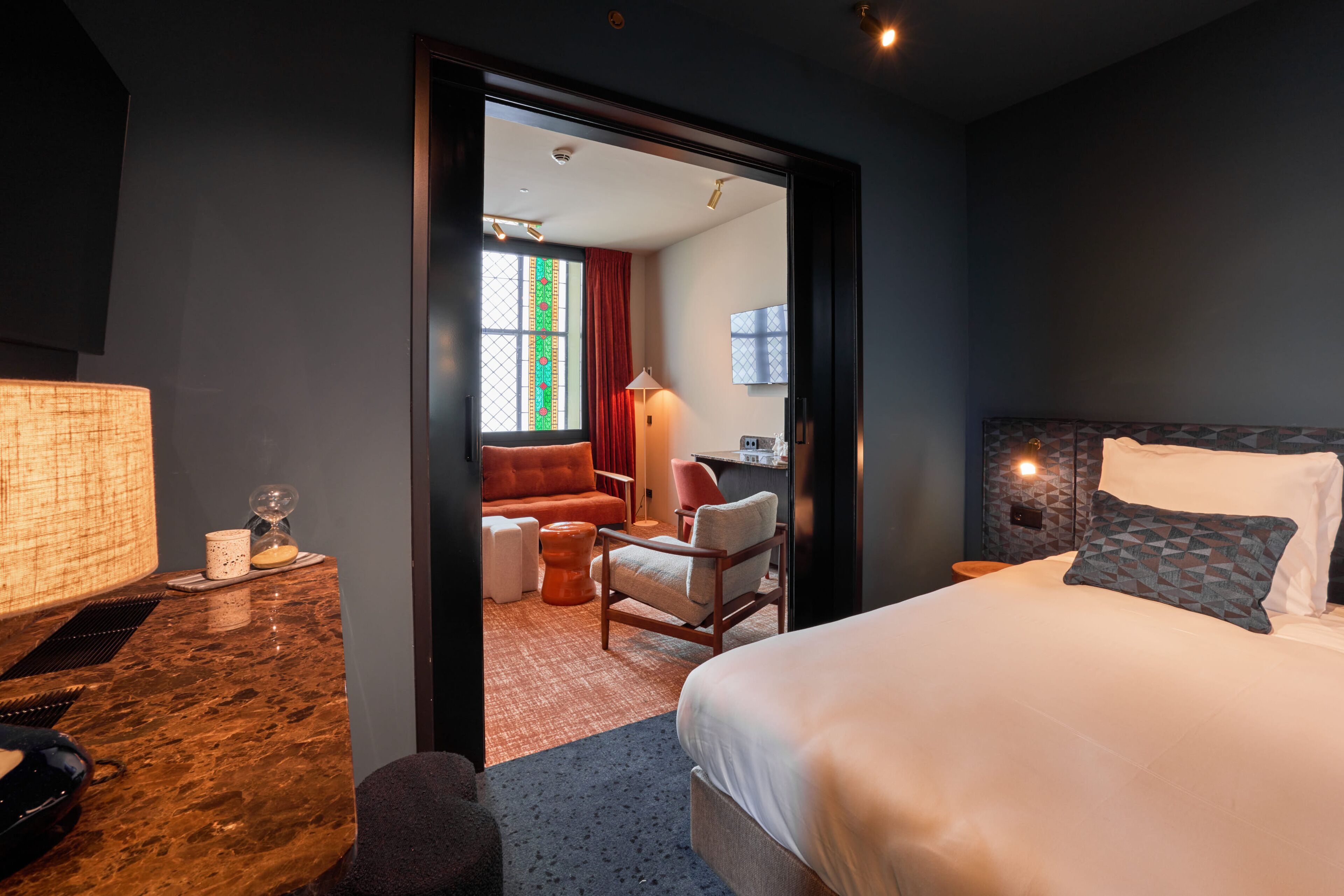 Une chambre d’hôtel avec un grand lit, un bureau avec une lampe, une chaise, une petite table sur laquelle se trouve un sablier, et une vue sur une autre chambre avec un canapé et des rideaux.