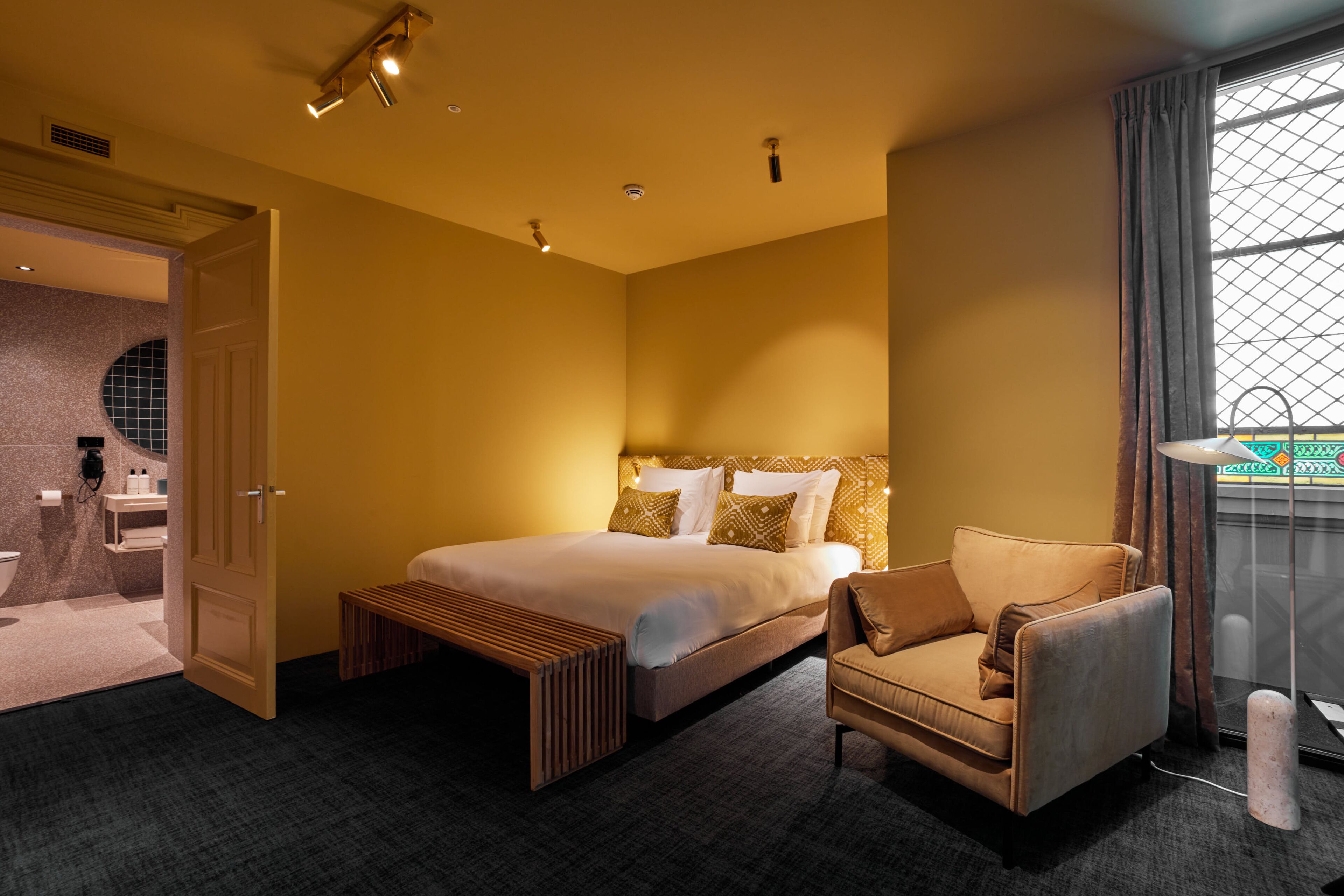 Una habitación de hotel con cama, una silla, un banco, una ventana con cortinas y un baño visible a través de una puerta abierta.
