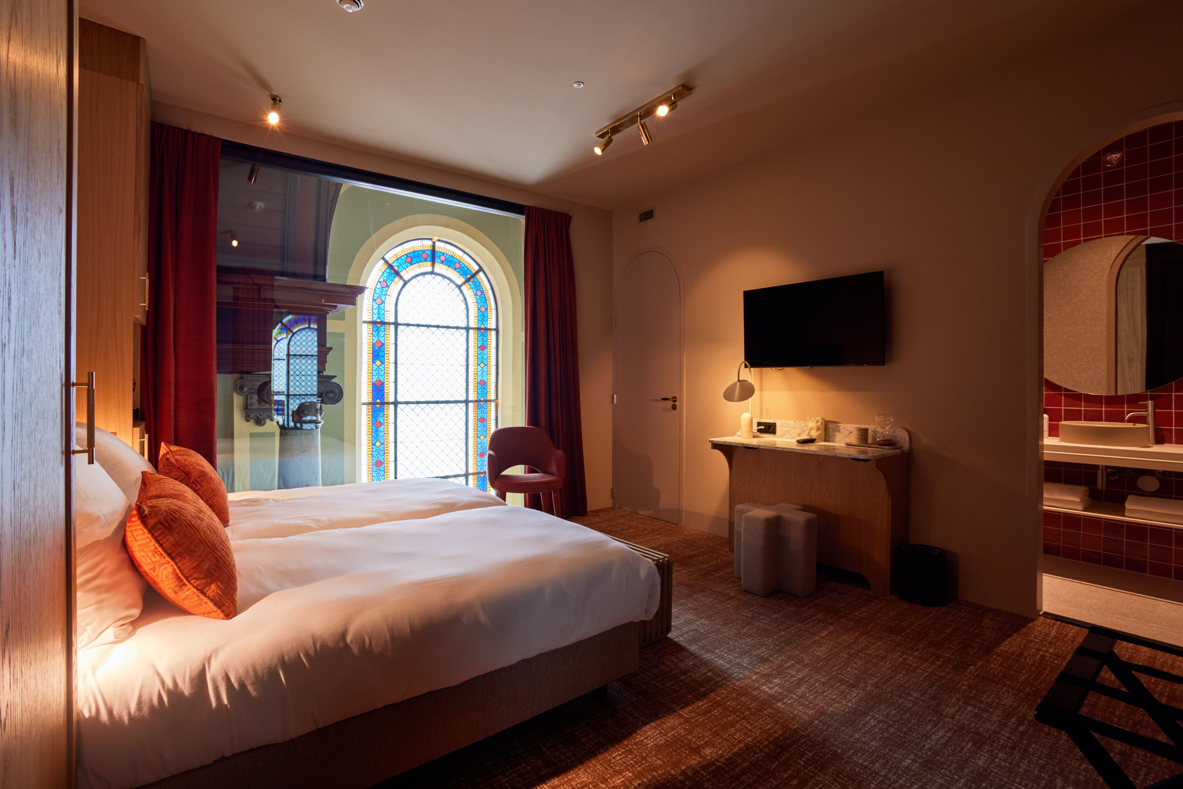Habitación de hotel con una cama grande, un escritorio, una silla, un televisor y un baño con pared de azulejos rojos.