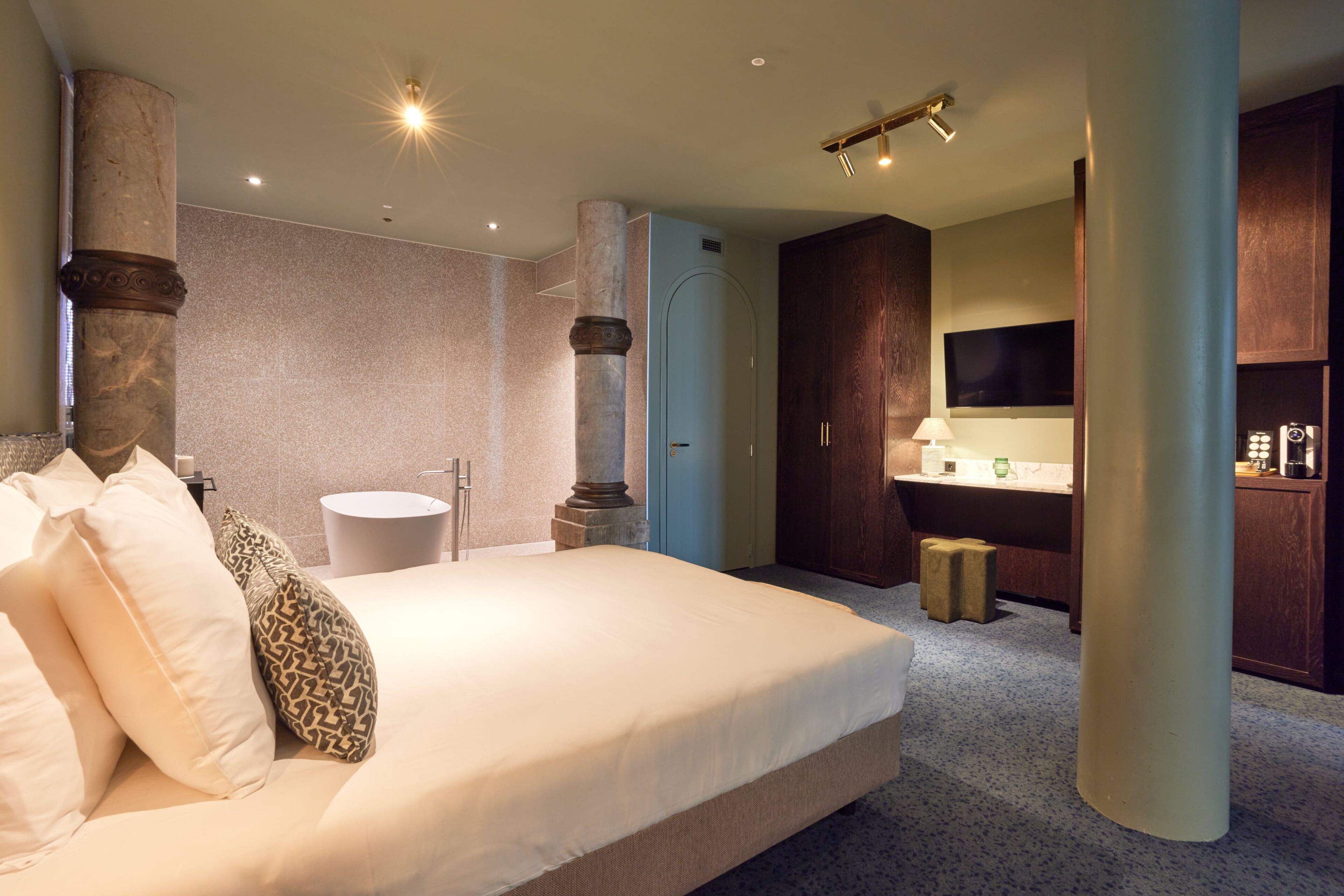 A modern hotel room with a large bed, a bathroom with a sink and mirror, and a TV mounted on the wall.