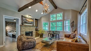 A spacious living room with a stone fireplace, a large sofa, a coffee table, and a dining area with a chandelier. - The Wildcat's Lair: Renovated Home in Highlands w/ Private Deck, Smart Home Tech (Highlands)