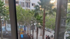 Vista a través de una ventana que muestra una calle con palmeras, coches estacionados y un edificio con ventanas arqueadas.