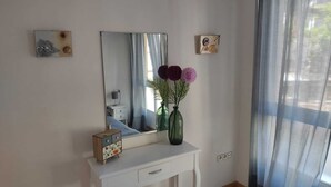 Una habitación con un escritorio blanco, un espejo, un jarrón con flores y una ventana con cortinas de gasa.