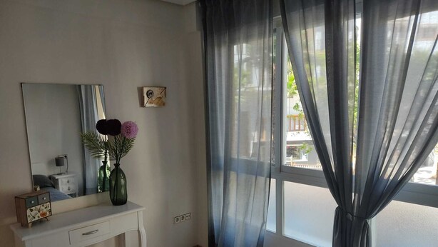 Un dormitorio con cama, mesita de noche con un jarrón de flores, cómoda, espejo y cortinas traslúcidas sobre una ventana.