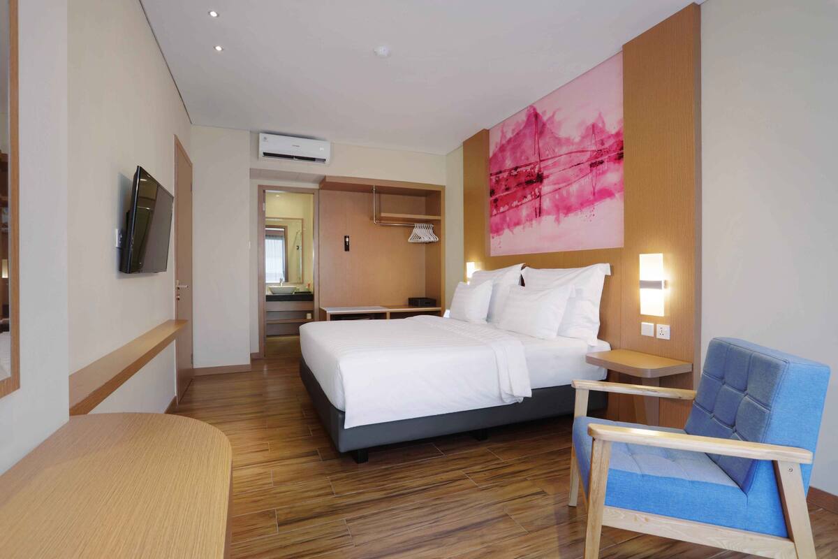 A modern hotel room with a large bed, a blue armchair, a wooden floor, a wall-mounted TV, and a large abstract painting.