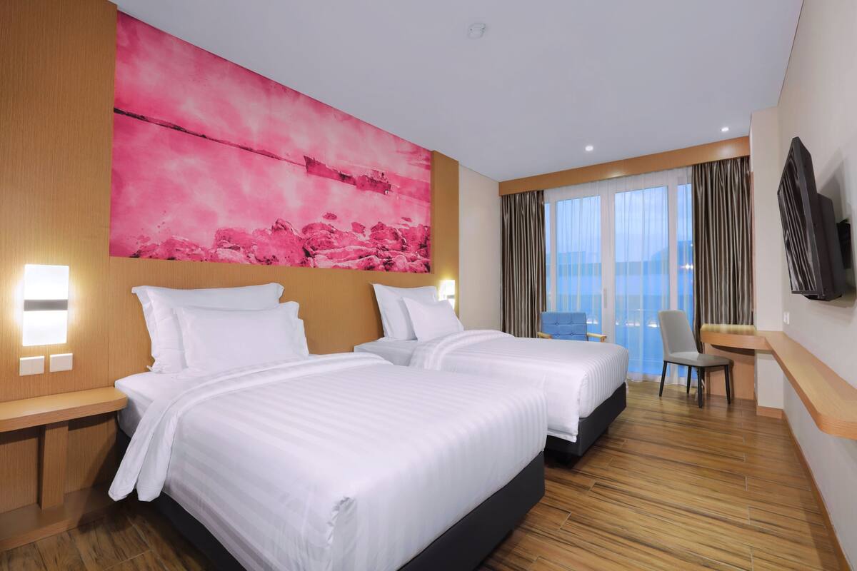 A hotel room with two beds, a large pink artwork on the wall, a TV mounted on the wall, and a view of the city through large windows.