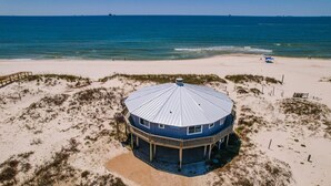 A beach house with a round blue roof, surrounded by sand dunes and a beach.