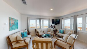 Uma sala de estar espaçosa com uma grande janela com vista para o mar, uma televisão fixada na parede e uma mesa de centro de madeira.