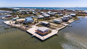 An aerial view of a coastal residential area with houses connected by piers to the water, a marina, and a beach in the background.