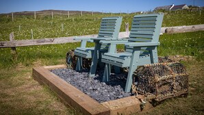 A rustic outdoor seating area with two green Adirondack chairs and a wooden table, surrounded by a stone border and a lobster trap. - The Byre, Waternish, Isle of Skye - Luxury Self Catering (Isle of Skye)