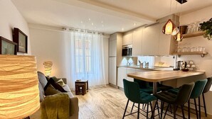 Una cocina moderna con una isla central, un comedor con sillas verdes y una zona de estar acogedora con una lámpara y cojines.