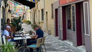 Una calle estrecha con mesas al aire libre en un café, un restaurante con toldo rojo y banderas triangulares coloridas.