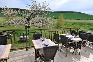 An outdoor dining area with tables and chairs, a metal railing, and a view of a green field and a tree with white blossoms.