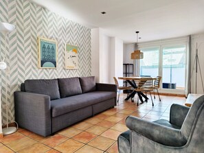 A living room with a grey sofa, a dining table with chairs, and a large window with a view.