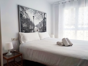 A neatly made bed with white linens and a rolled towel, a bedside table with a lamp, and a large black and white cityscape painting on the wall.