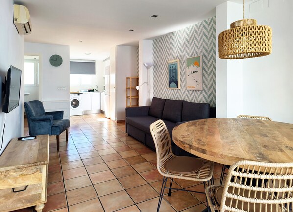 A modern living space with a sofa, dining table, wicker chairs, a TV, and a kitchen in the background.