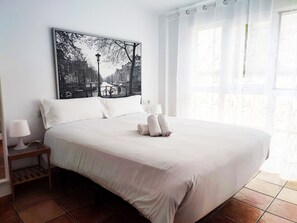 A neatly made bed with white linens and towels, a nightstand with a lamp, and a large black-and-white cityscape painting on the wall.