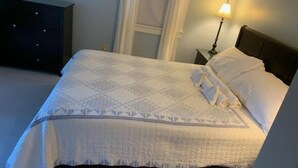 A neatly made bed with a white quilt and pillows, a bedside lamp, and a dark dresser in a room with light-colored walls. - Oak Bluffs Rental - Walk to the Beach (Oak Bluffs)