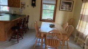 A dining area with a table and chairs, a kitchen area with wooden cabinets, and a window with blinds. - Oak Bluffs Rental - Walk to the Beach (Oak Bluffs)