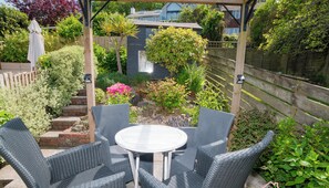 A patio with wicker furniture and a small table, surrounded by a garden with various plants and a wooden fence. - Stylish Coastal 3 Bed Haven | Close to town on Coronation Road (Salcombe)