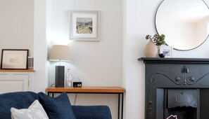 A living room with a blue sofa, a small table with a lamp, a framed picture on the wall, and a fireplace with a vase and plant. - Stylish Coastal 3 Bed Haven | Close to town on Coronation Road (Salcombe)