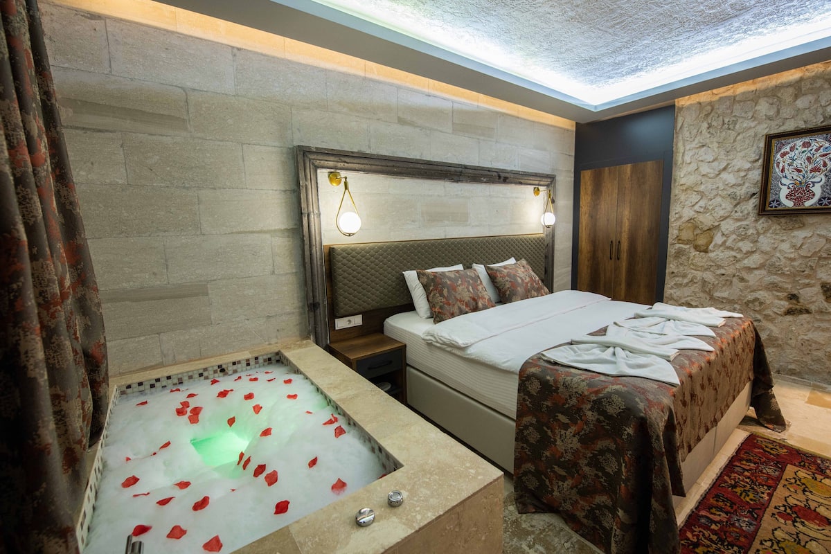 A bedroom with a large bed, a nightstand, a wall-mounted lamp, a stone wall, and a bathtub with rose petals.