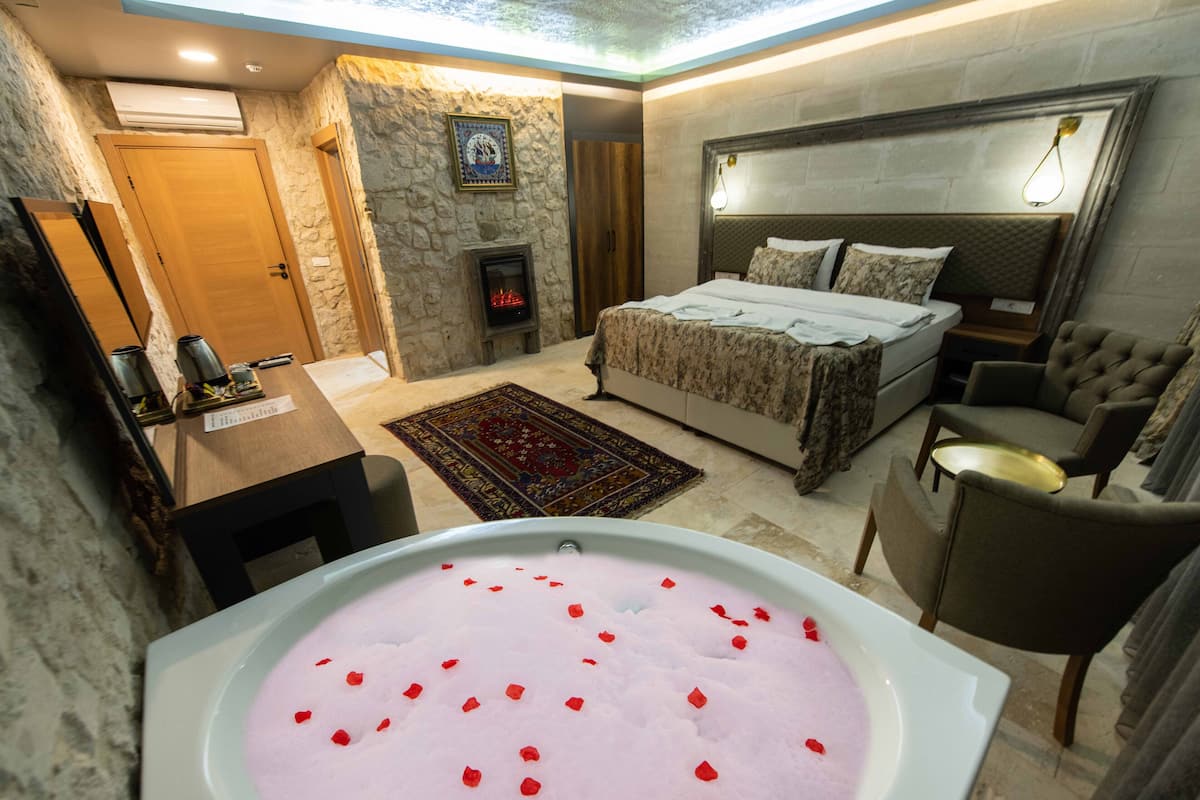A bedroom with a stone wall, a bed, a nightstand, a chair, a desk, a fireplace, and a bathtub with rose petals.