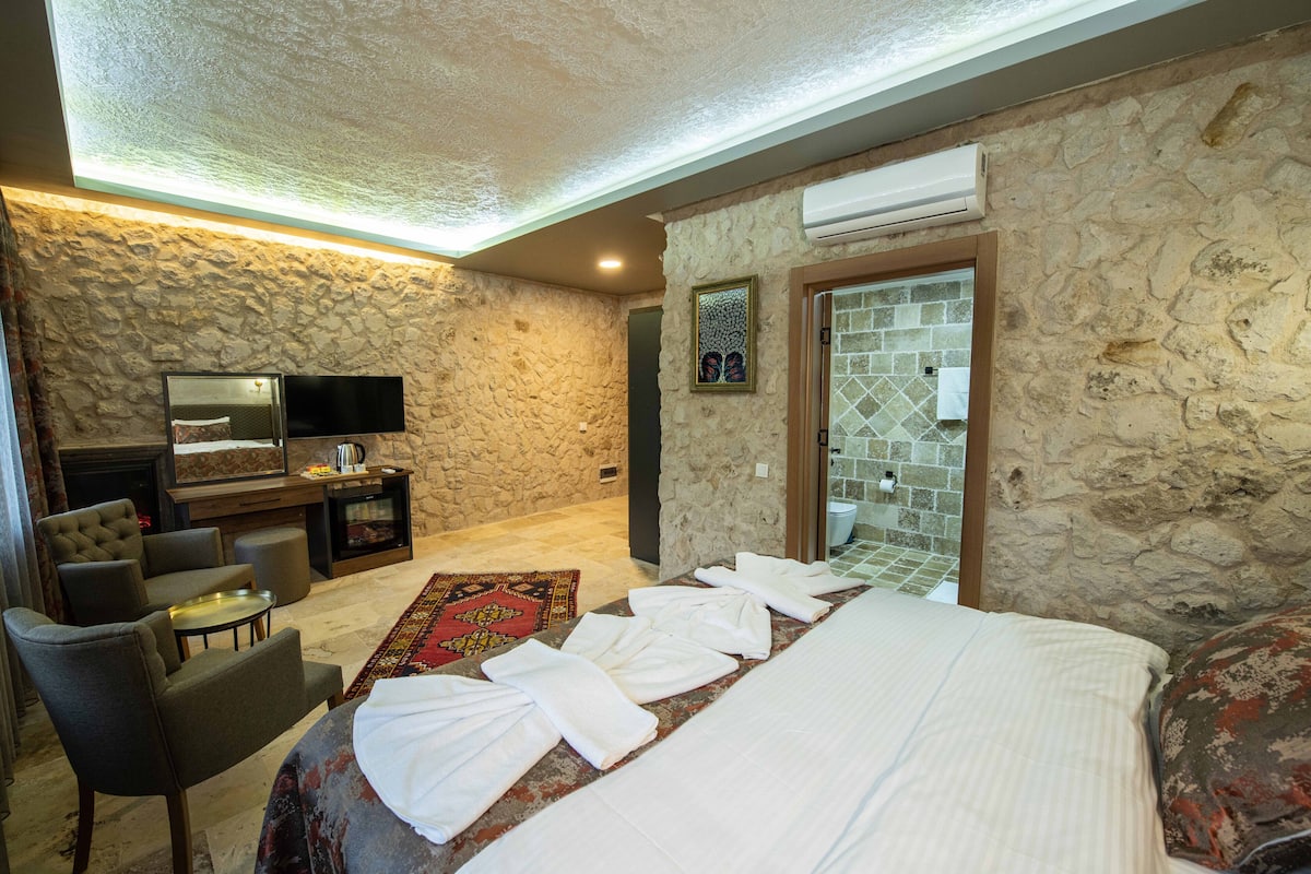 A hotel room with a stone wall, a flat-screen TV, a sofa, a coffee table, a bed with towels, and a bathroom with a tiled shower.