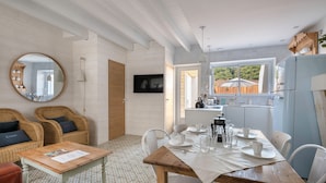 Ein Essbereich mit einem Holztisch, gedeckt für vier Personen, umgeben von Korbstühlen. Ein Fernseher ist an der Wand befestigt, und ein Fenster bietet einen Blick nach draußen.