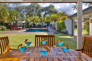 A wooden table with four blue cocktails garnished with orange slices, set outdoors with a view of a swimming pool and tropical trees.